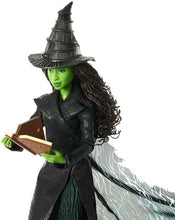 Action figure of a witch character with green skin, black hat, and long hair, holding a book.
